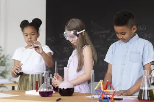 Three children conduct a science experiment at a lab table, with colorful liquids in glassware; girl in safety goggles stands center, others helping nearby.